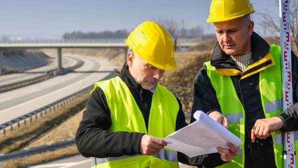 Two civil engineers wearing hard hats and safety vests refer to paper documents near a freeway.