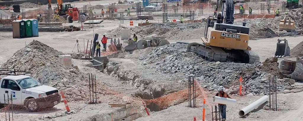 Trucks, earth-moving equipment, and construction workers move about an active construction site among rebar and aggregate.